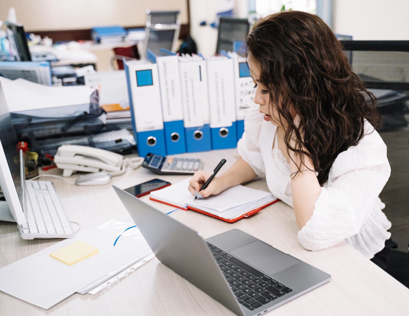 An office worker taking notes at a desk beside a laptop and documents