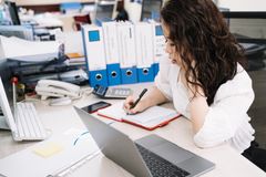 An office worker takes notes while working at a desk with a laptop and documents.