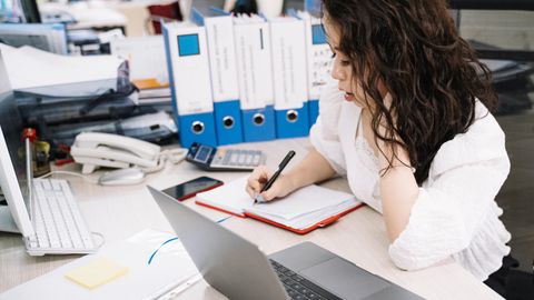 An office worker takes notes while working at a desk with a laptop and documents.