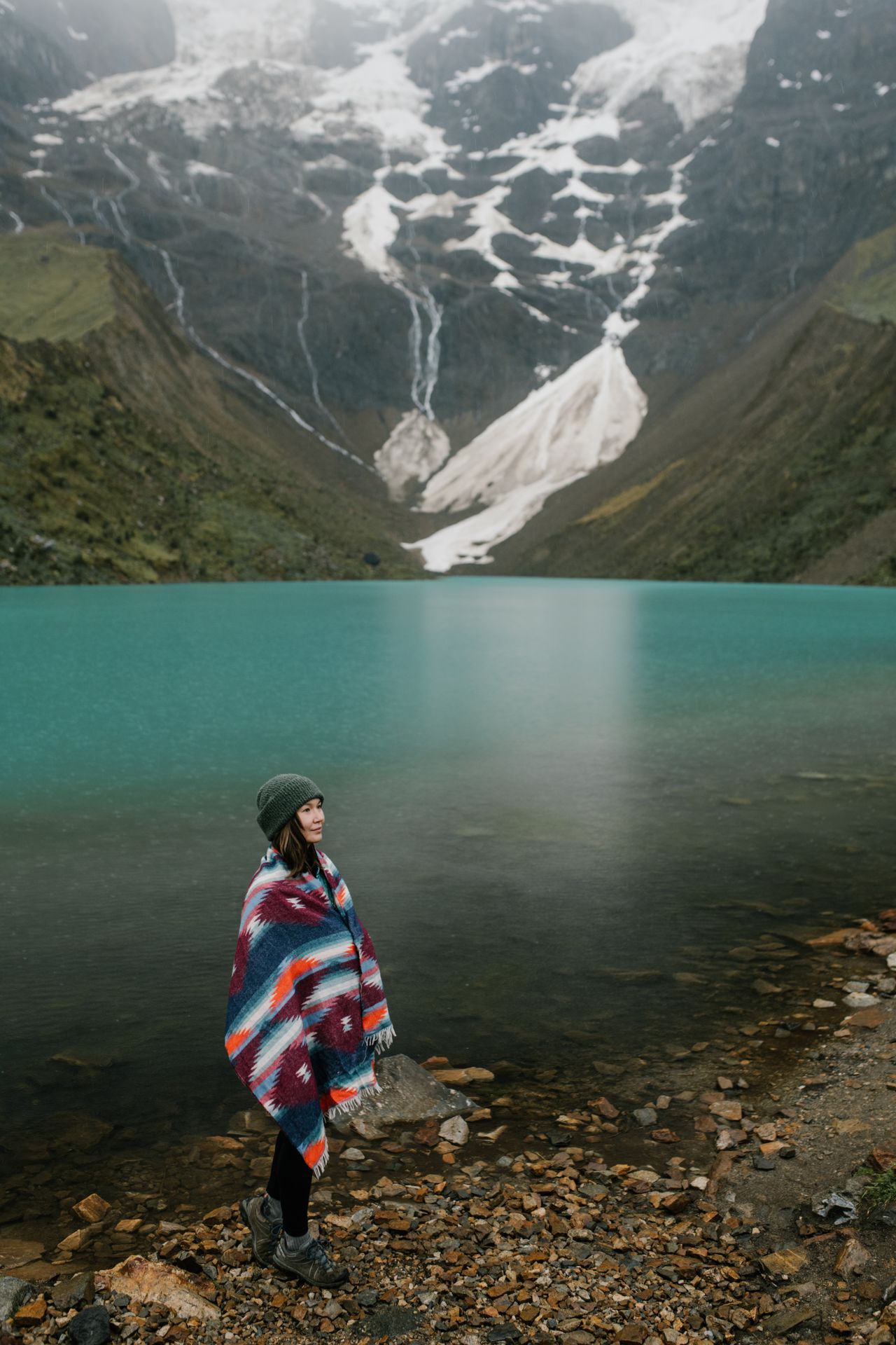 Woman wrapped in a colorful striped shawl by a mountain lake