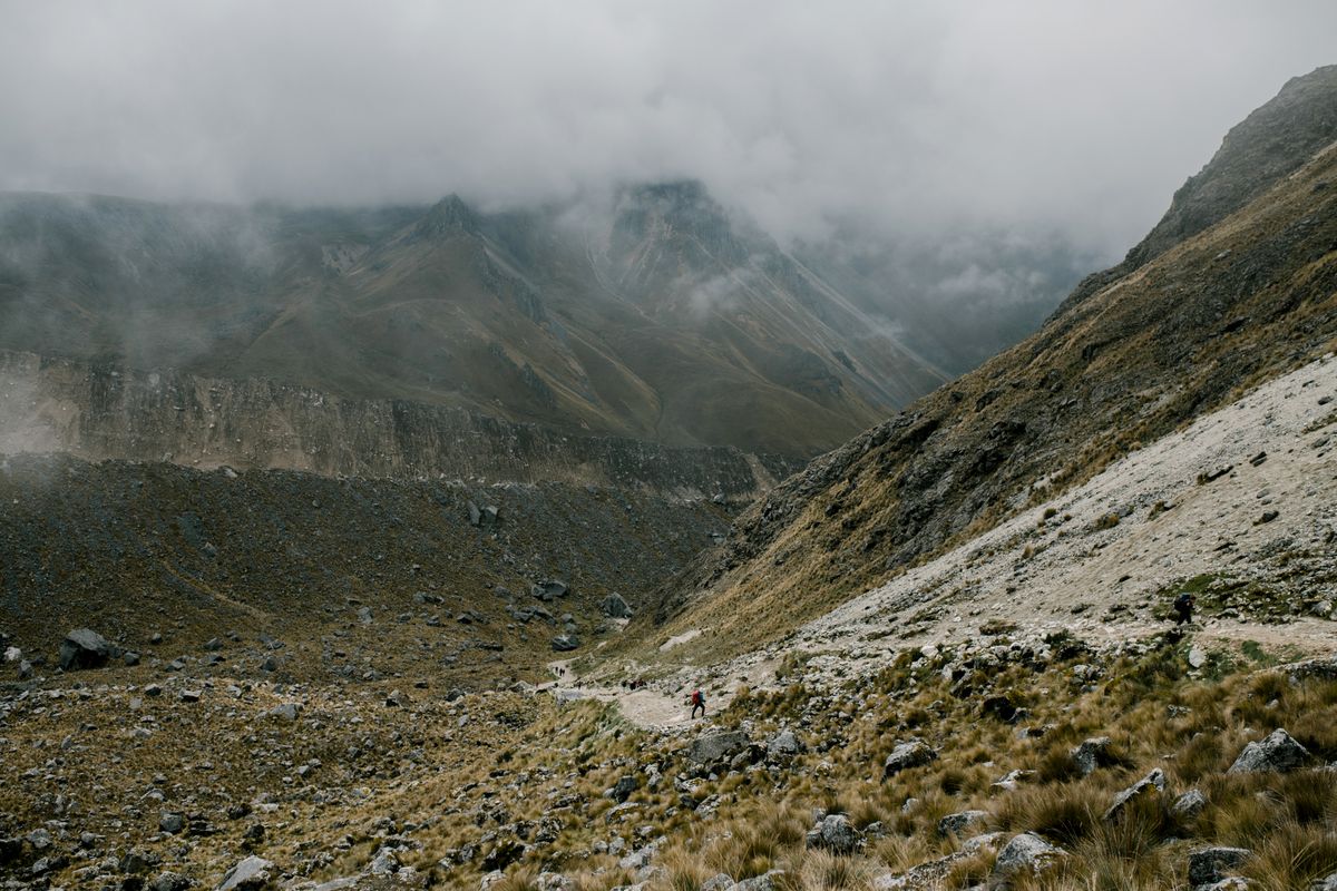 A misty green Andean valley descending below a rocky pass, a lone hiker visible far below on the trail, low cloud clinging to the peaks