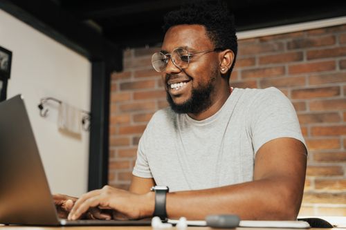 Free A man with glasses and beard smiling while typing on a laptop indoors. Stock Photo