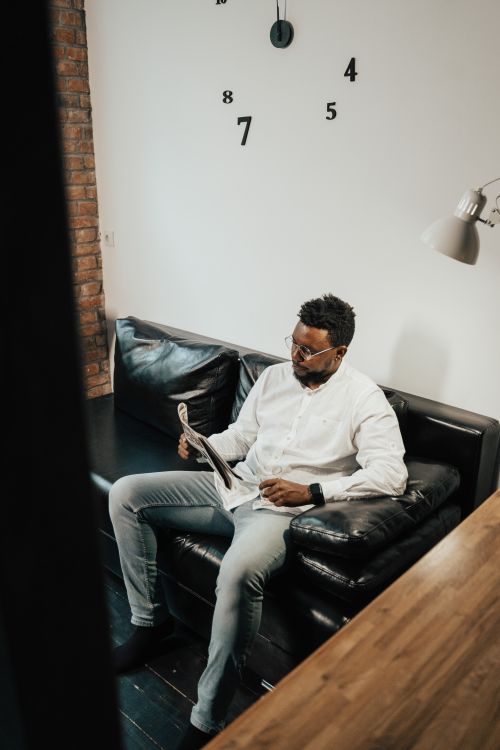 Free Man sitting on a black leather sofa reading a newspaper in a cozy, modern interior. Stock Photo