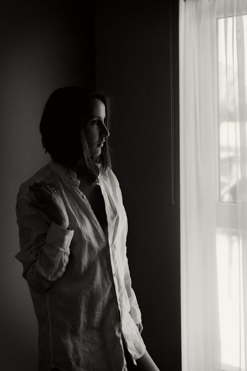 Black and white serene female wearing domestic shirt standing in room and looking out of window