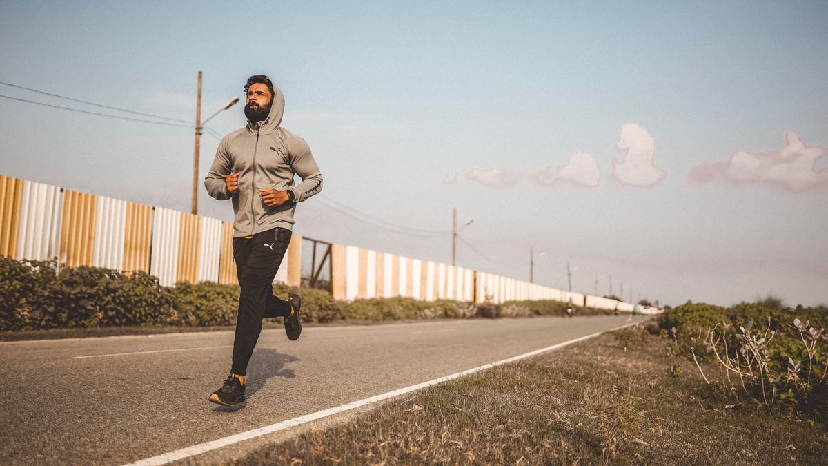 A bearded man in a hoodie jogs along an empty road under a clear sky.