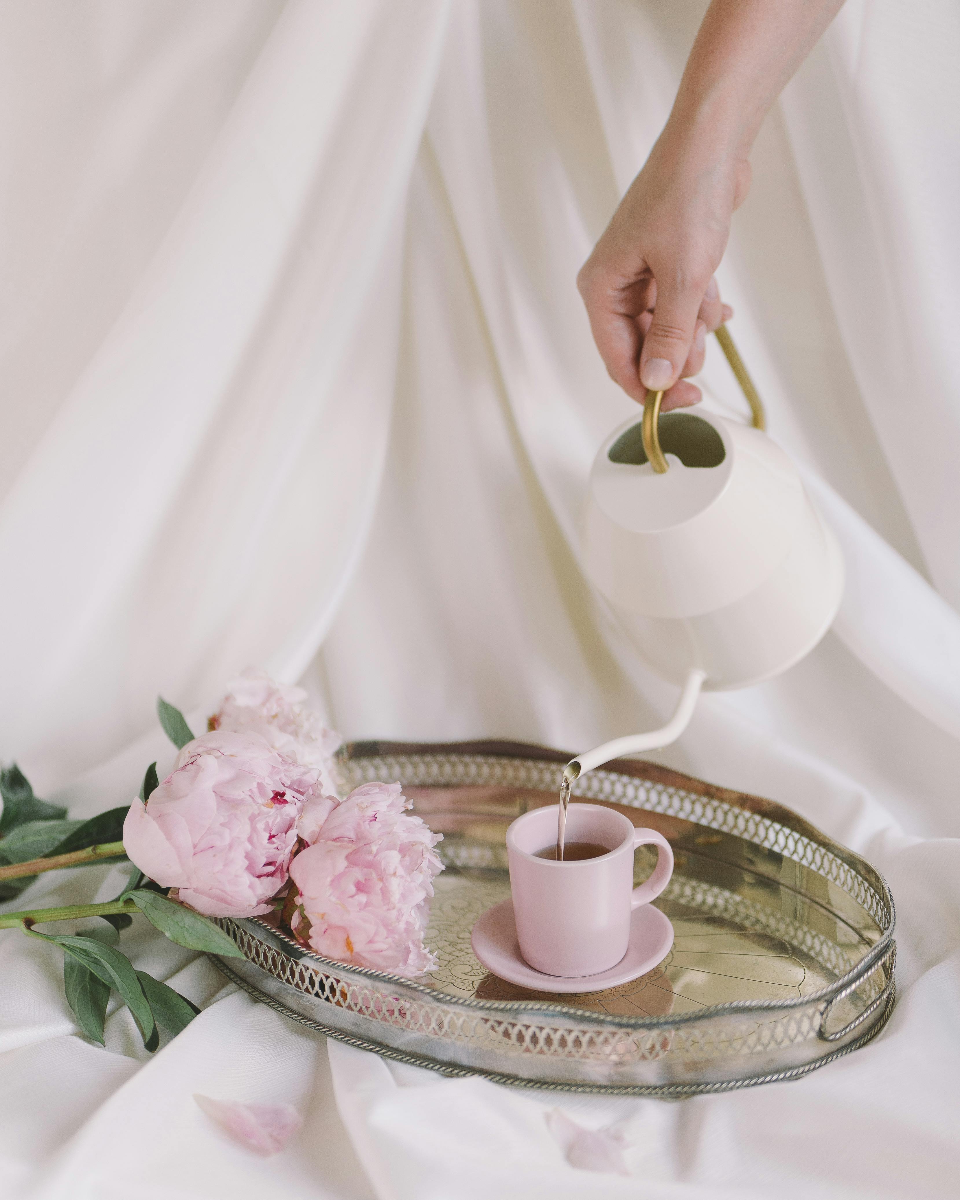 A hand pours tea into a cup on a tray with pink peonies. Elegant and serene setting.