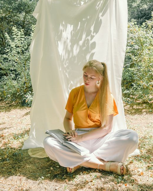 A woman in a yellow top and white pants enjoys reading a book outside.