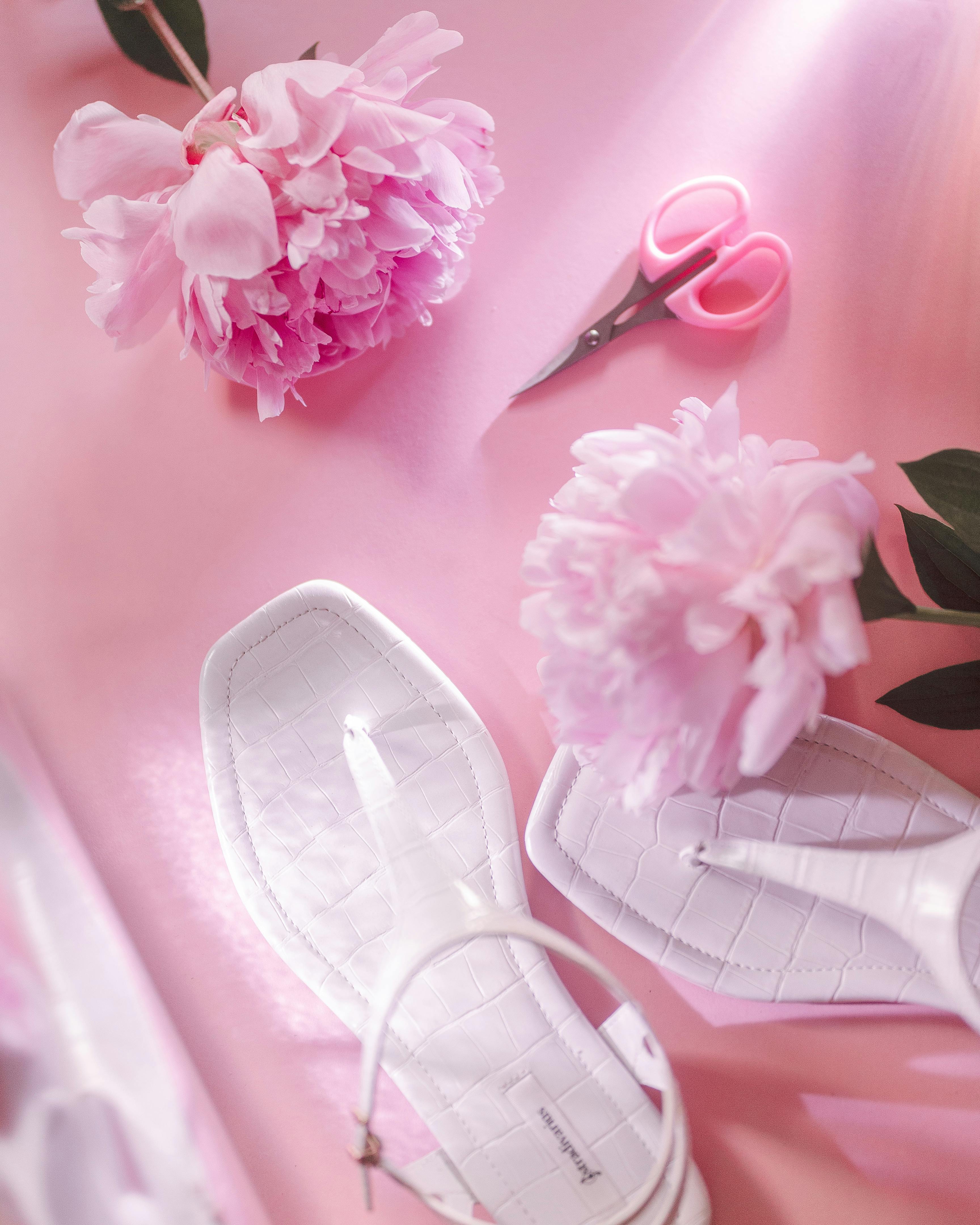 Chic arrangement with white sandals and vibrant pink peonies on a pastel pink background.