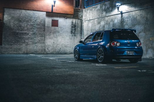 Blue hatchback automobile with spoiler parked on asphalt with marking lines near stone barrier in city street in evening time