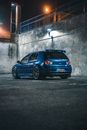 Blue hatchback automobile placed near cement wall with fence on parking space with marking in city street in night time