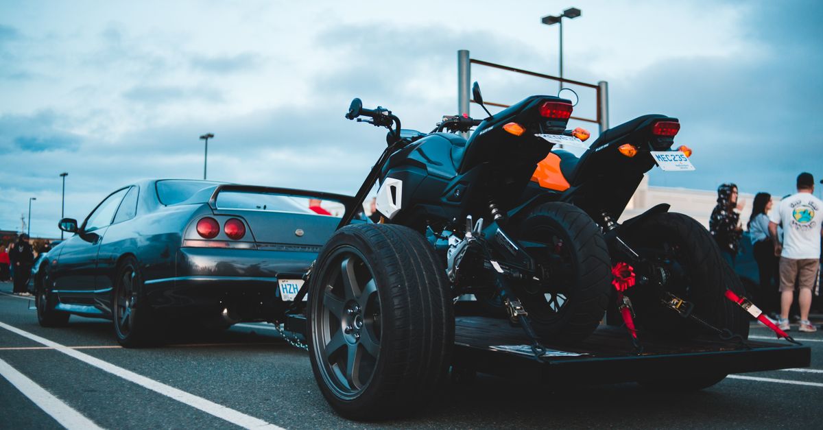 Black car towing trailer with modern motorcycles on asphalt road against cloudy sky in city street with distant people on roadside