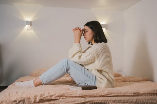 Free Woman in White Sweater Sitting and Praying on Bed Stock Photo