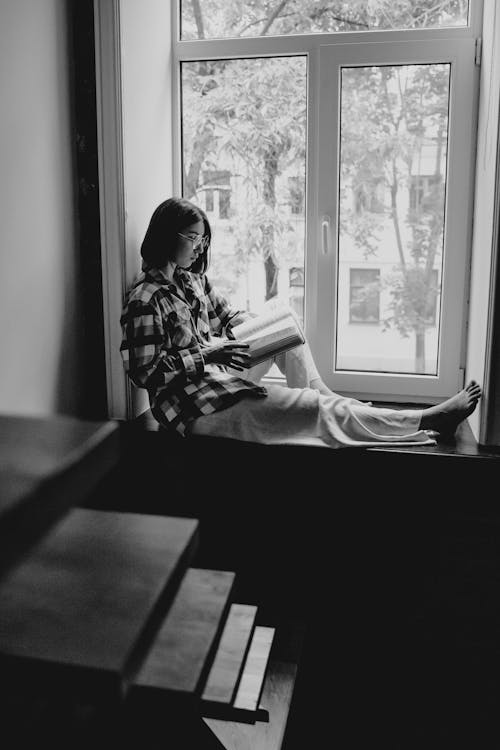 Grayscale Photo of Woman Reading a Book beside the Window
