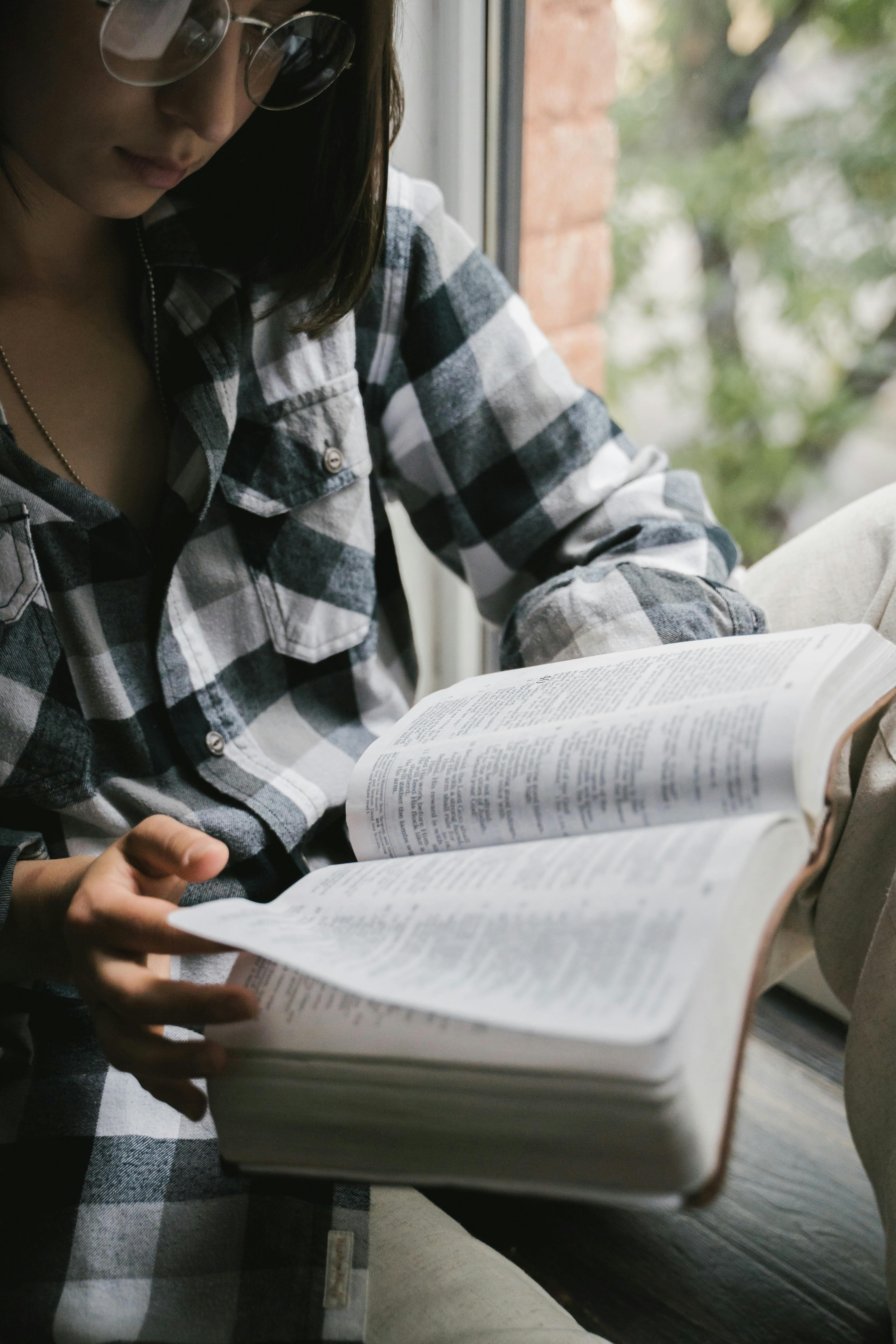Free A woman wearing glasses reads an open book by the window, in a plaid shirt. Relaxed indoor setting. Stock Photo