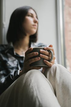 A young woman sits by a window holding a hot coffee mug, lost in thought.