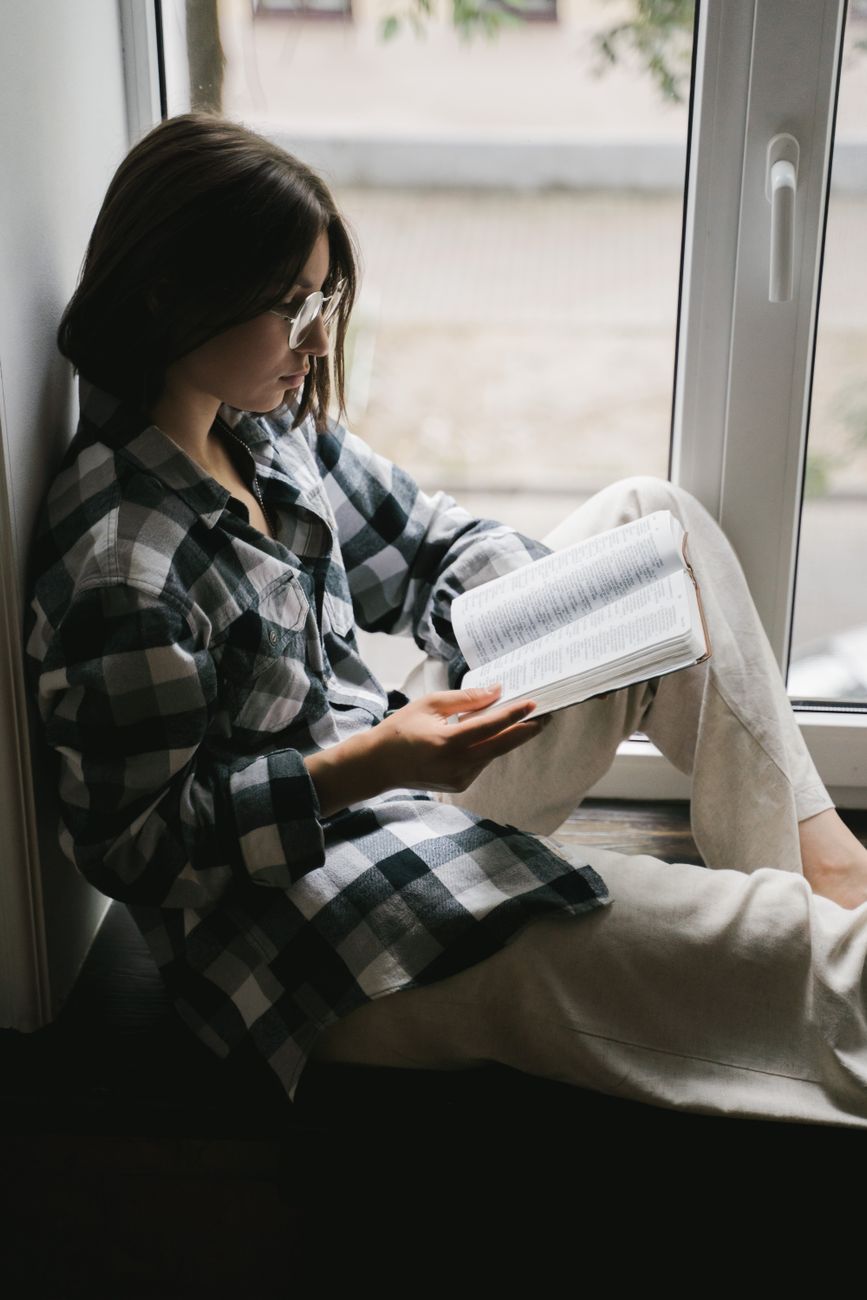 Young woman in glasses reading a book by the window, enjoying a quiet moment indoors.. pexels, young, woman