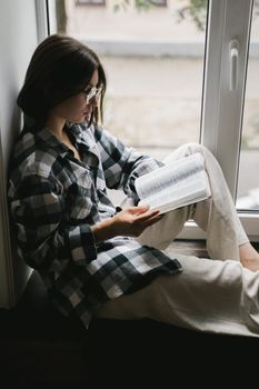 Young woman in glasses reading a book by the window, enjoying a quiet moment indoors.. pexels, young, woman