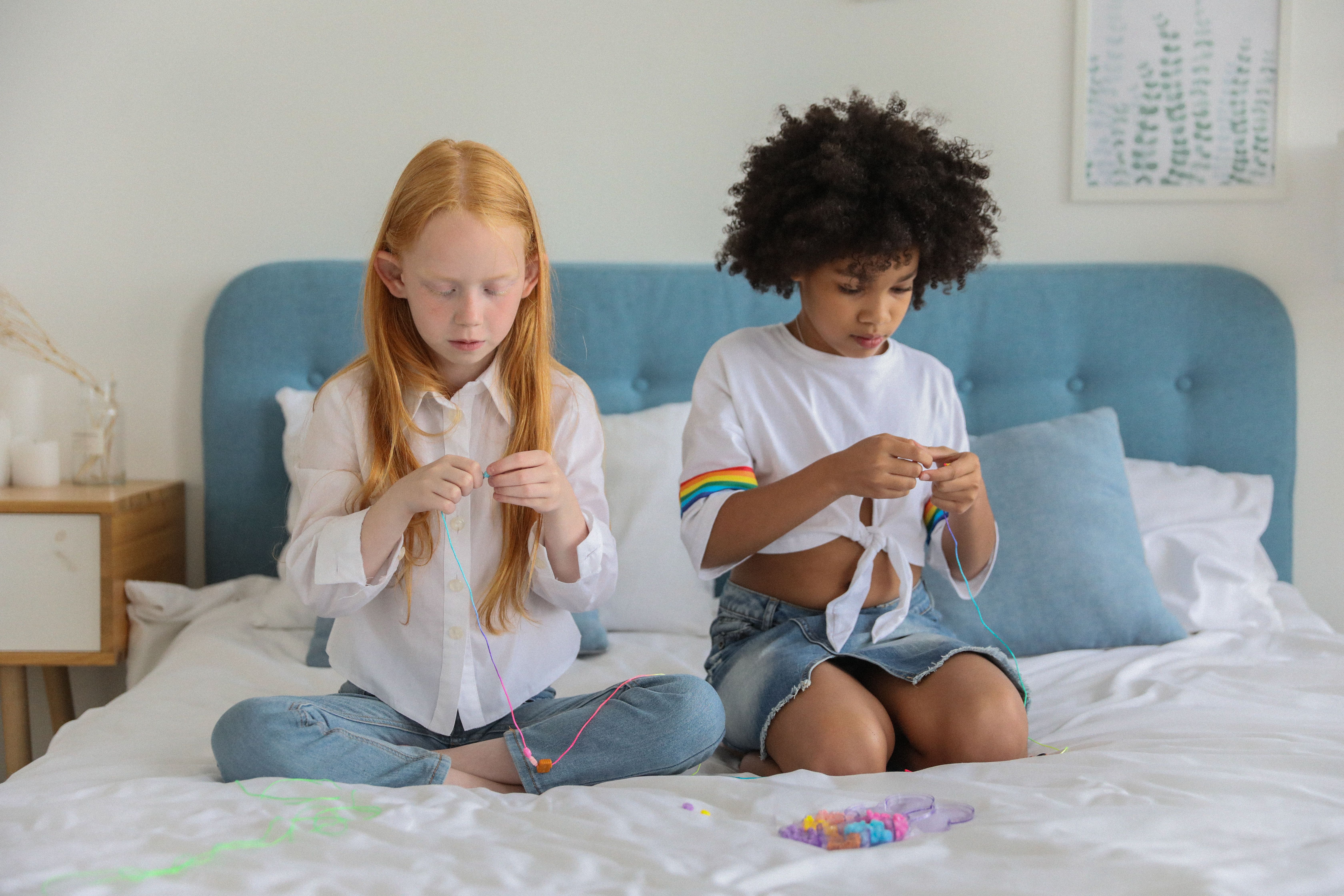 Two children crafting friendship bracelets on a bed in a bright, cozy room.
