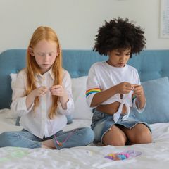 Two young children sitting on a bed threading beads onto string, a pile of beads between them