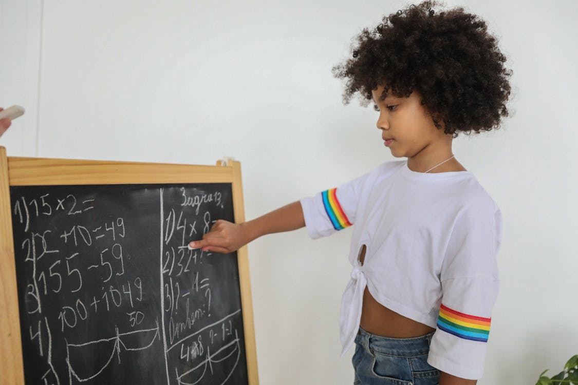 Side view of pensive black girl pointing at mathematical example on blackboard at light room in daytime