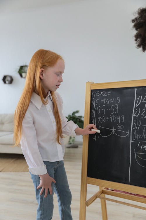 Concentrated schoolgirl standing near easel blackboard and writing mathematical equation in cozy light room
