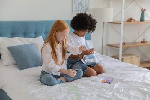 Multiethnic children stringing beads on thread while sitting with crossed legs on bed blanket at home
