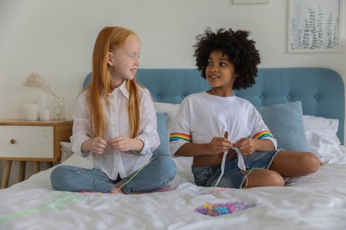 Cheerful multiracial best friends stringing beads on threads while looking at each other on bed blanket in house