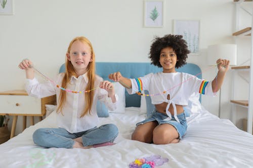 Glad multiracial girlfriends showing beads on threads while sitting on soft bed and looking at camera at home