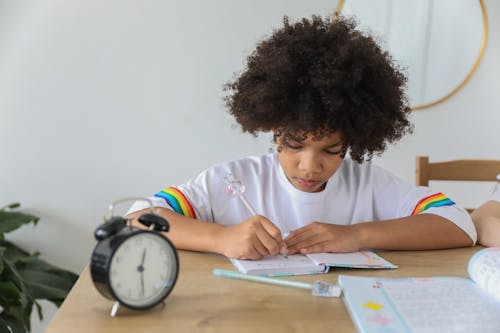 Concentrated African American child writing in notebook while studying at desk with alarm clock at home