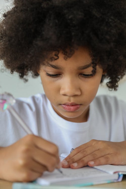 Crop attentive African American schoolgirl taking note in diary while studying at desk in daylight