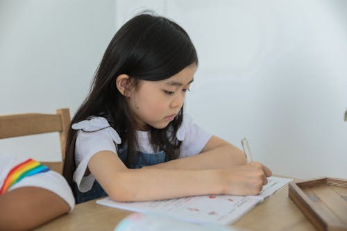 Concentrated ethnic schoolkid writing in activity book with pen while studying at table near unrecognizable black friend