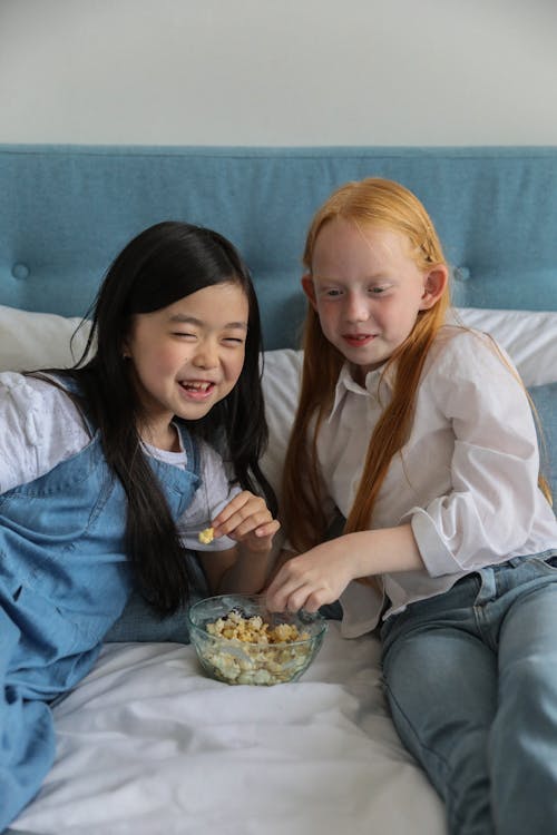 Happy Asian kid with best friend eating popcorn while spending time together on soft bed and looking away