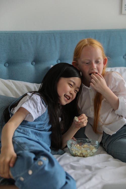 Happy diverse girlfriends eating popcorn on bed at home