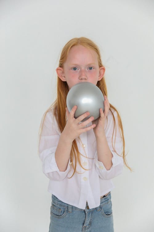 Child with red head blowing shiny grey balloon while looking at camera on white background
