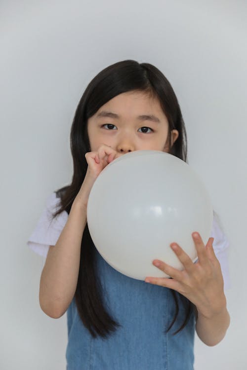 Adorable little Asian girl with long dark hair in casual clothes inflating balloon and looking at camera against white background