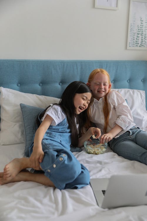 Cute multiracial children eating popcorn while lying on bed and watching funny movie on laptop