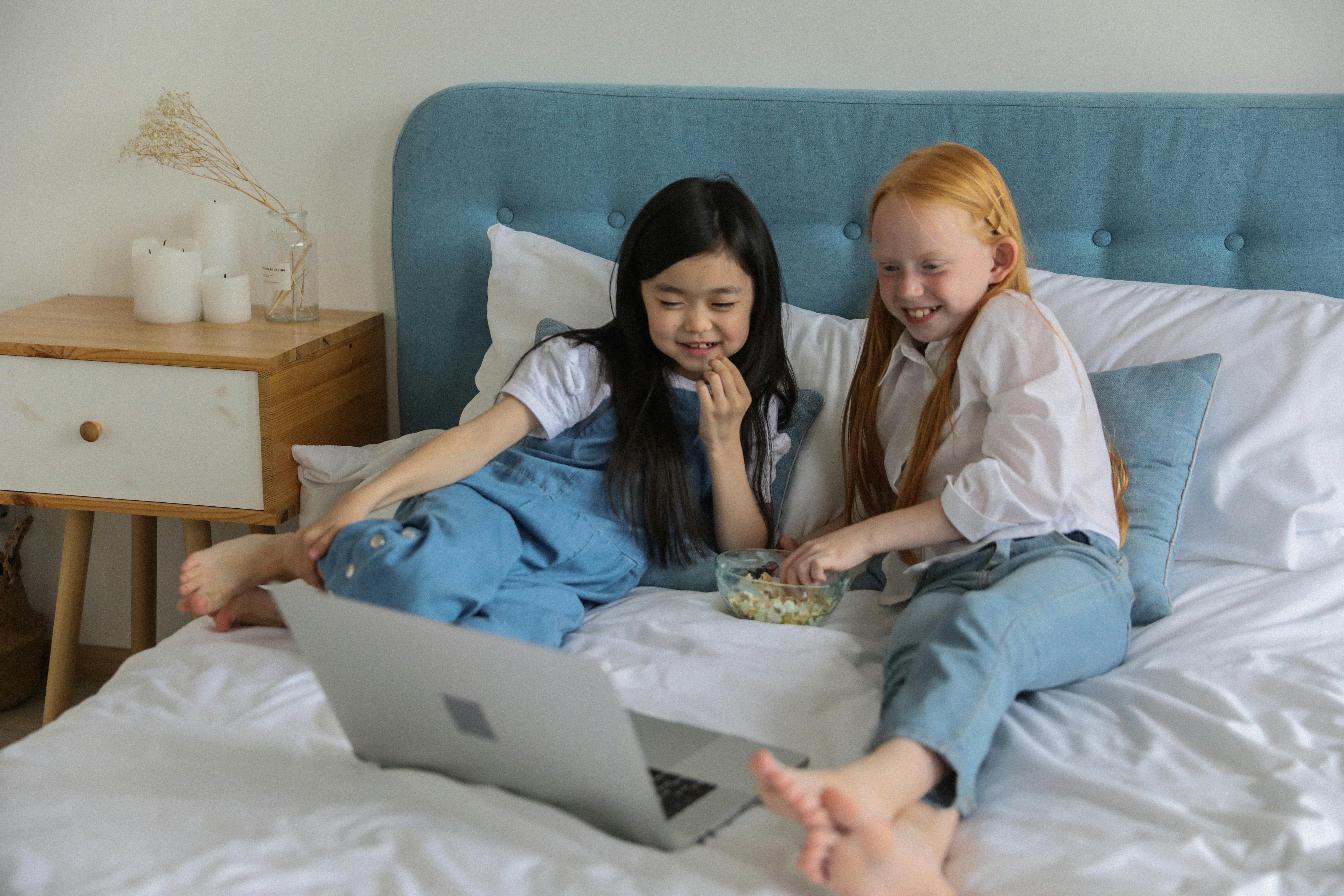 cheerful little multiethnic girlfriends watching movie on laptop and eating popcorn on bed