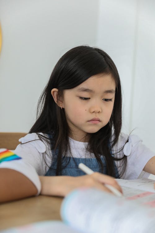 Free Attentive cute little Asian girl with long dark hair sitting at table and writing in copybook during lesson at school Stock Photo