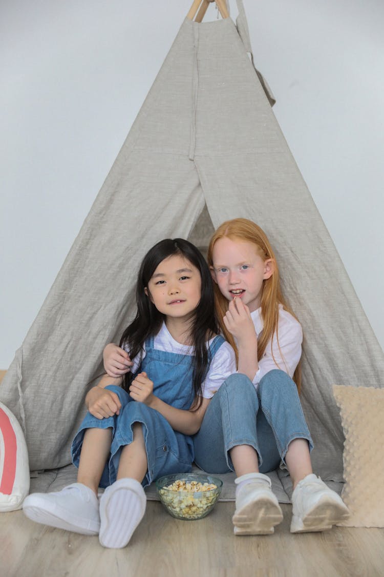 Happy Little Diverse Children Hugging And Eating Popcorn While Playing Together In Tent At Home