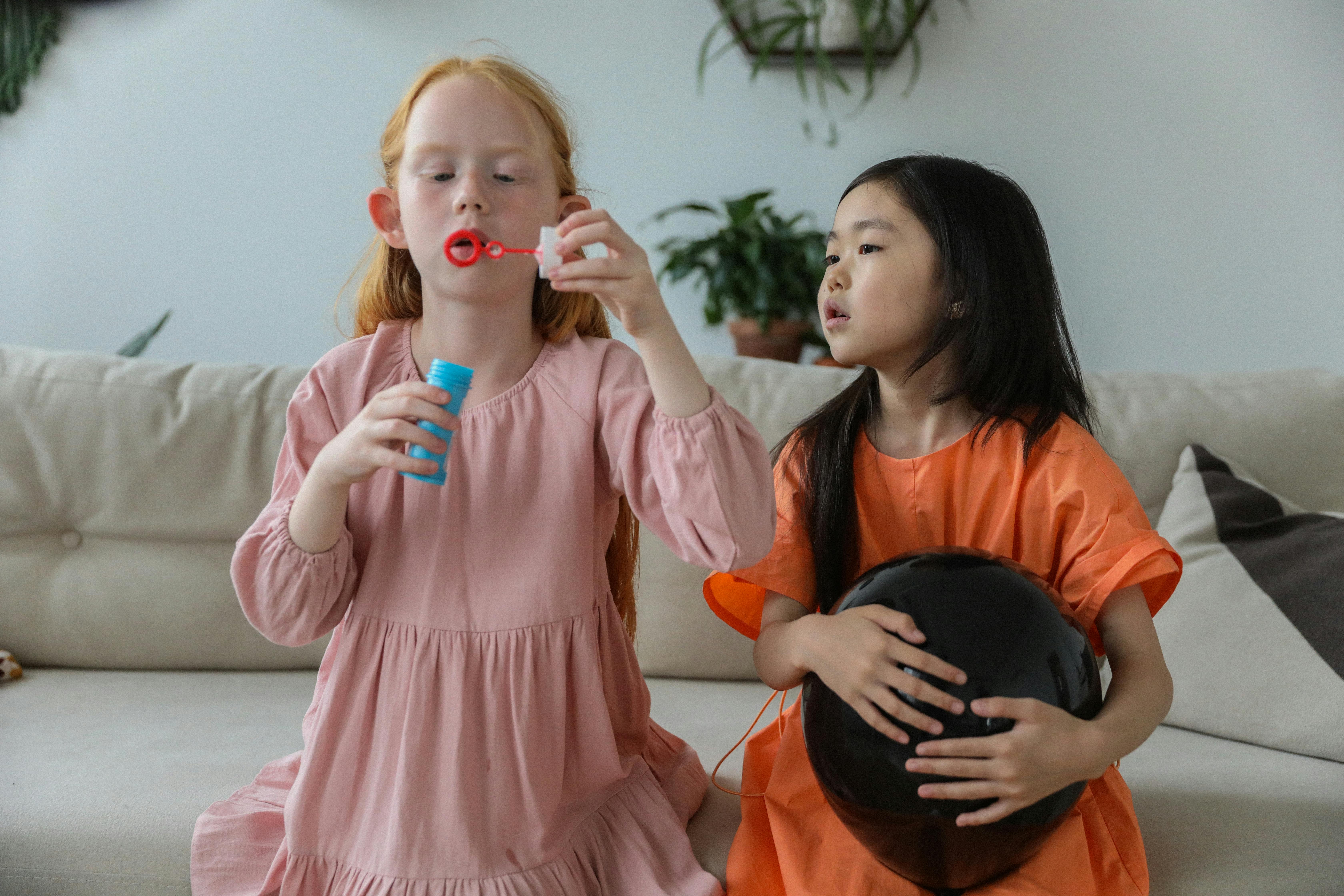 cute little diverse girls in trendy dresses blowing soap bubbles while playing together on couch