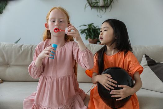 Adorable little redhead girl in stylish dress blowing soap bubbles while playing with cute Asian girlfriend holding black balloon sitting on sofa at home