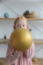 Adorable child in girlish dress inflating balloon at home