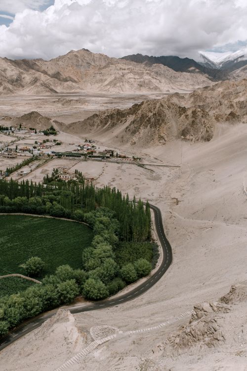 Free Drone view of narrow road going among green trees and desert with mountains with settlement in distance against cloudy sky in nature Stock Photo