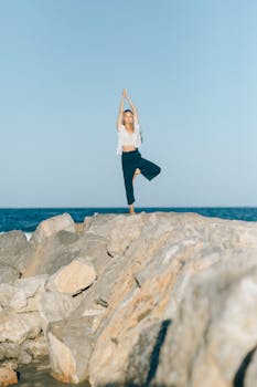 Woman practicing yoga in tree pose on rocky seashore under clear sky.