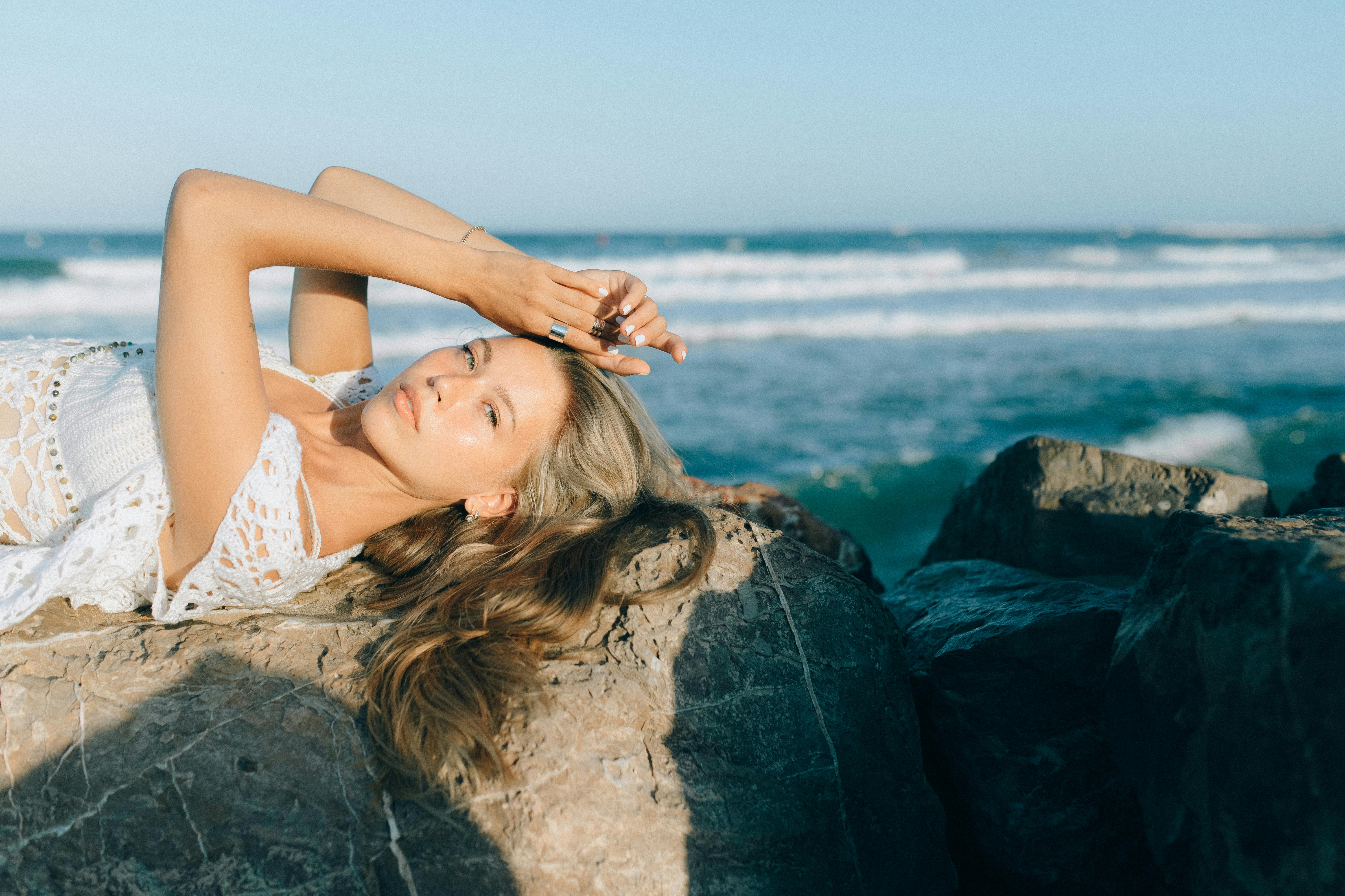阳光明媚的一天，一位身着波西米亚服饰的年轻女子正悠闲地坐在海边的岩石上