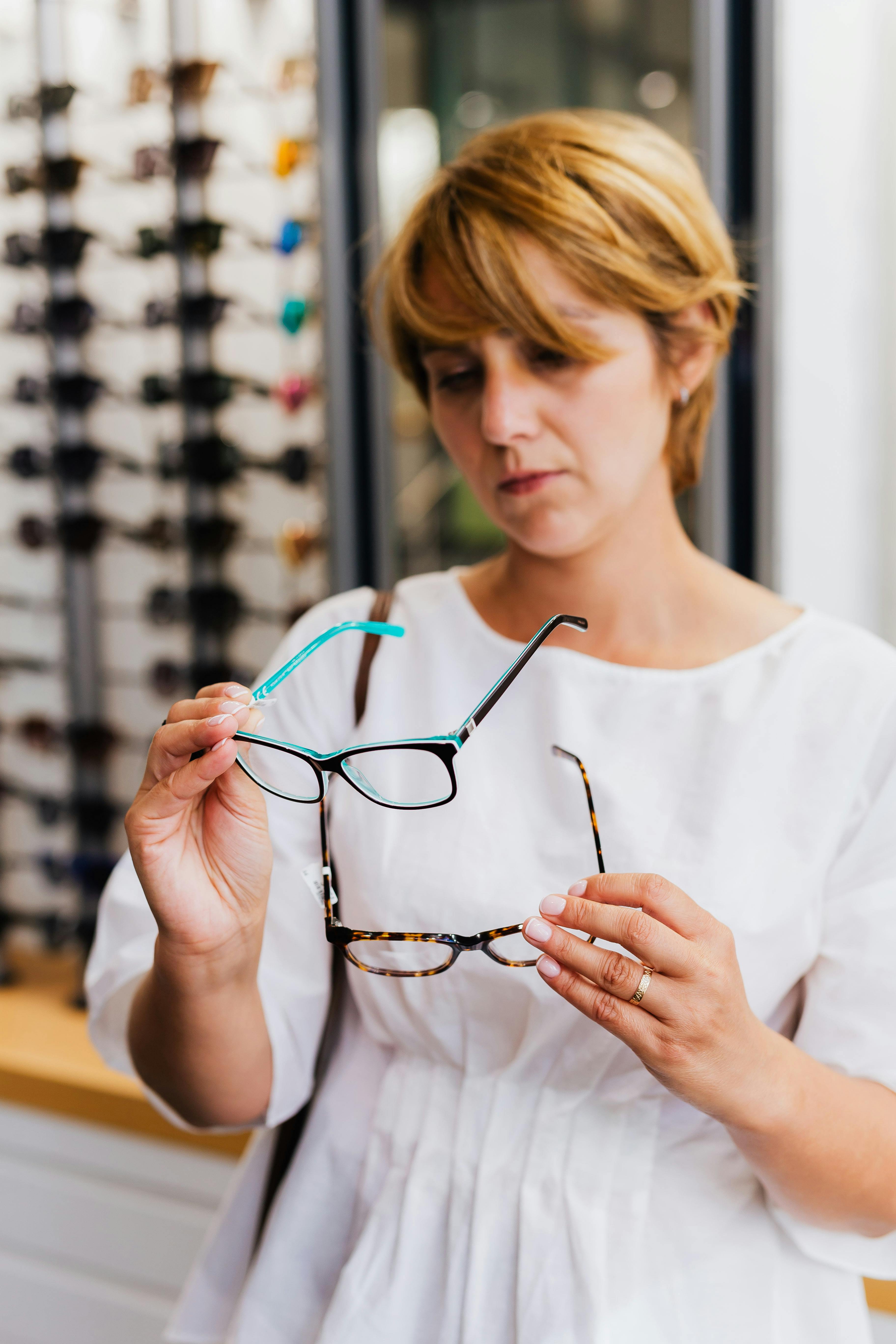 A woman examining eyeglasses options in an optical store, wearing a white dress.