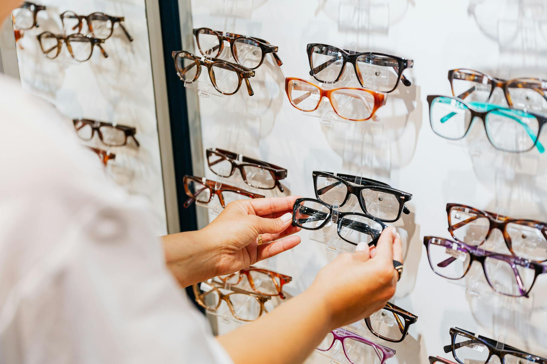 Hands selecting glasses from a display wall in an eyewear store.