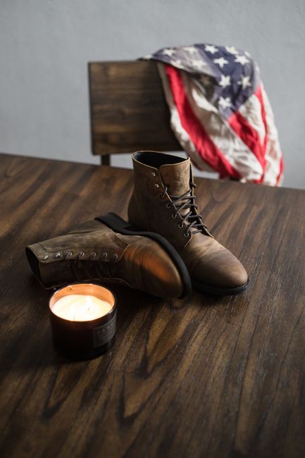 Leather boots on wooden table with candle and American flag backdrop, evoking patriotism.