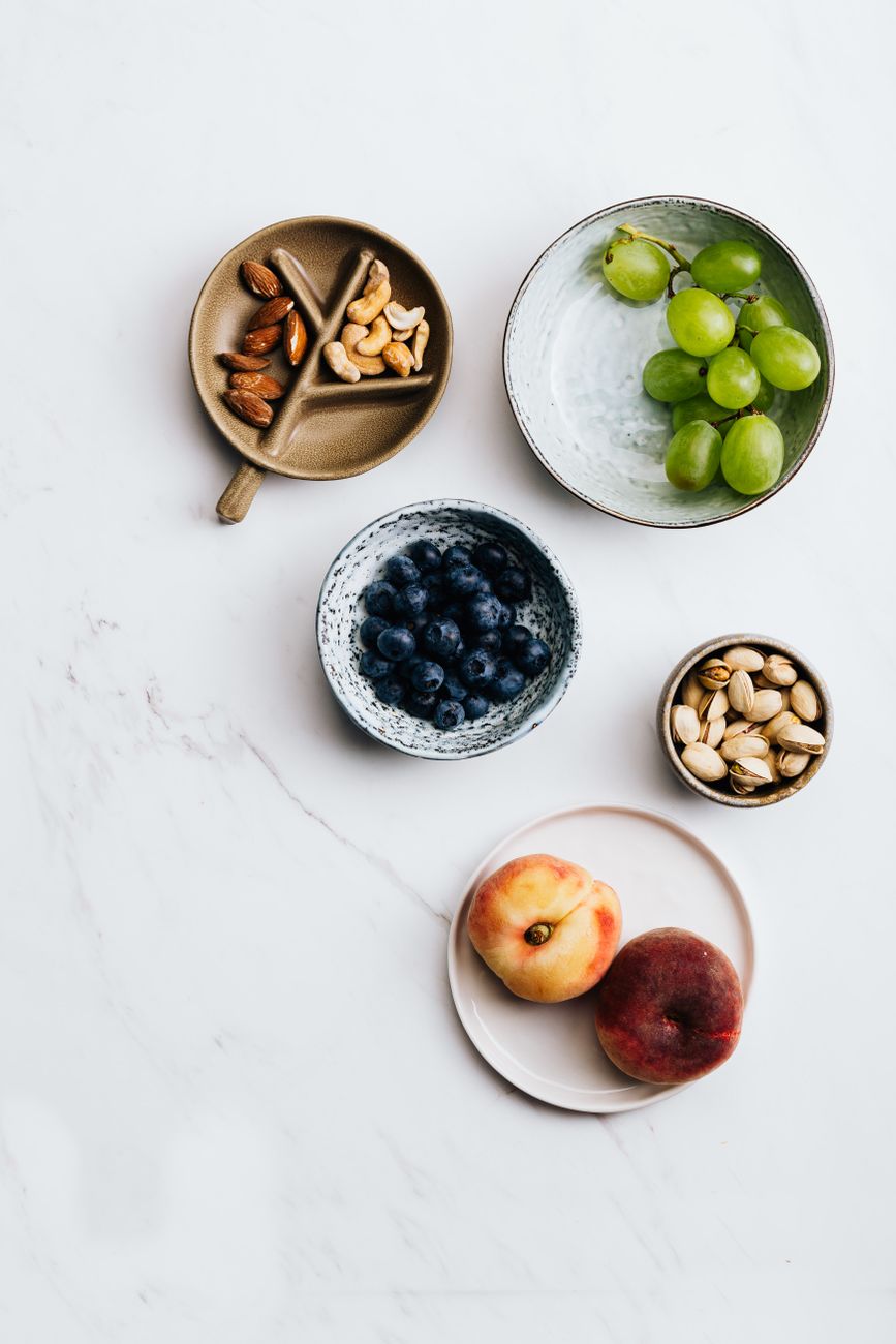 Flat lay of fresh fruits and nuts in bowls on a white marble surface, perfect for health and lifestyle themes.