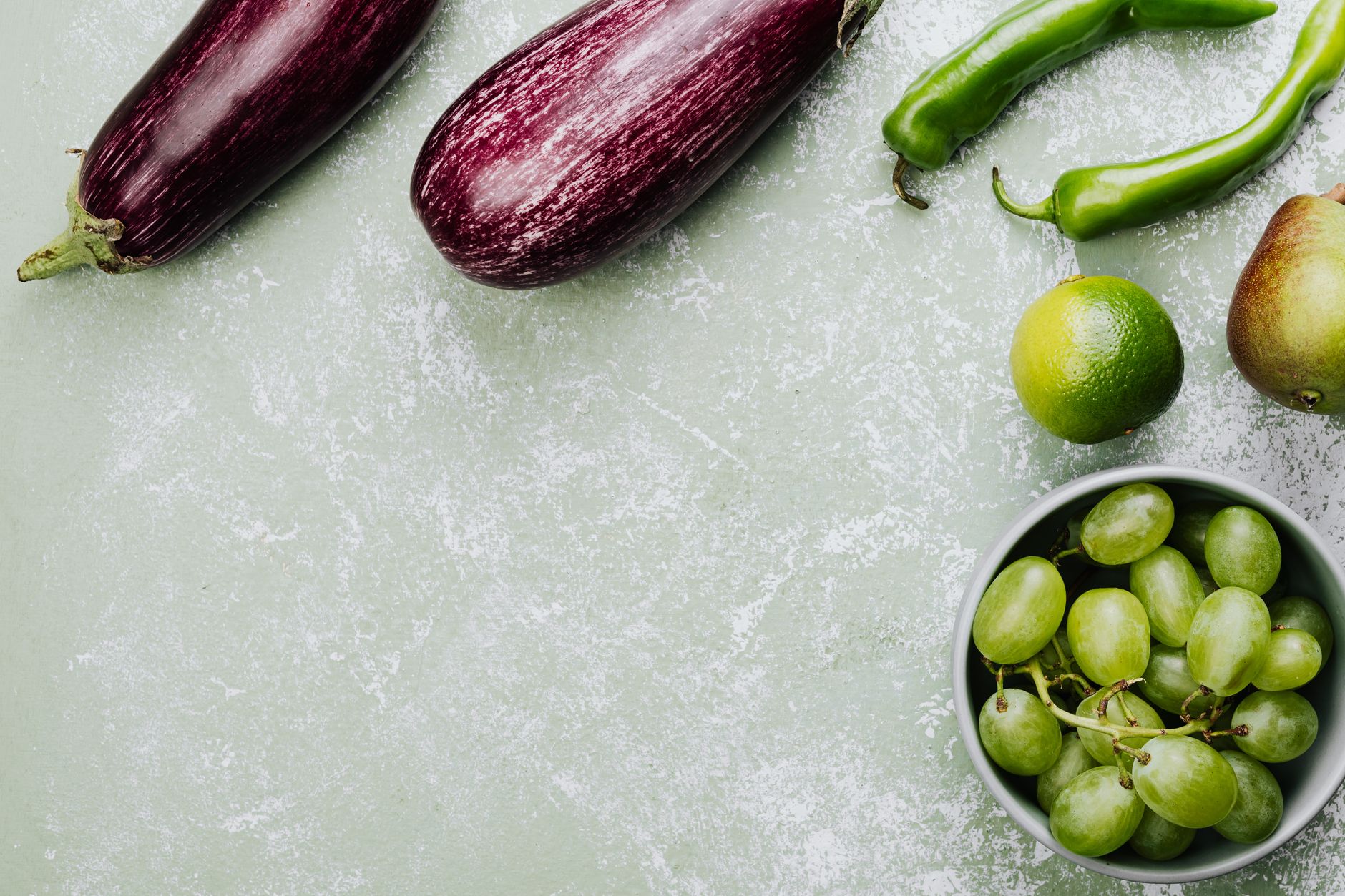 Top view of fresh vegetables and fruits featuring eggplants, green peppers, lime, pear, and grapes on a green surface.